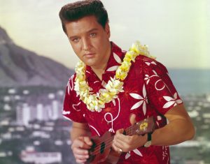 Elvis Presley wearing in an aloha shirt, flower lei, while playing ukulele.