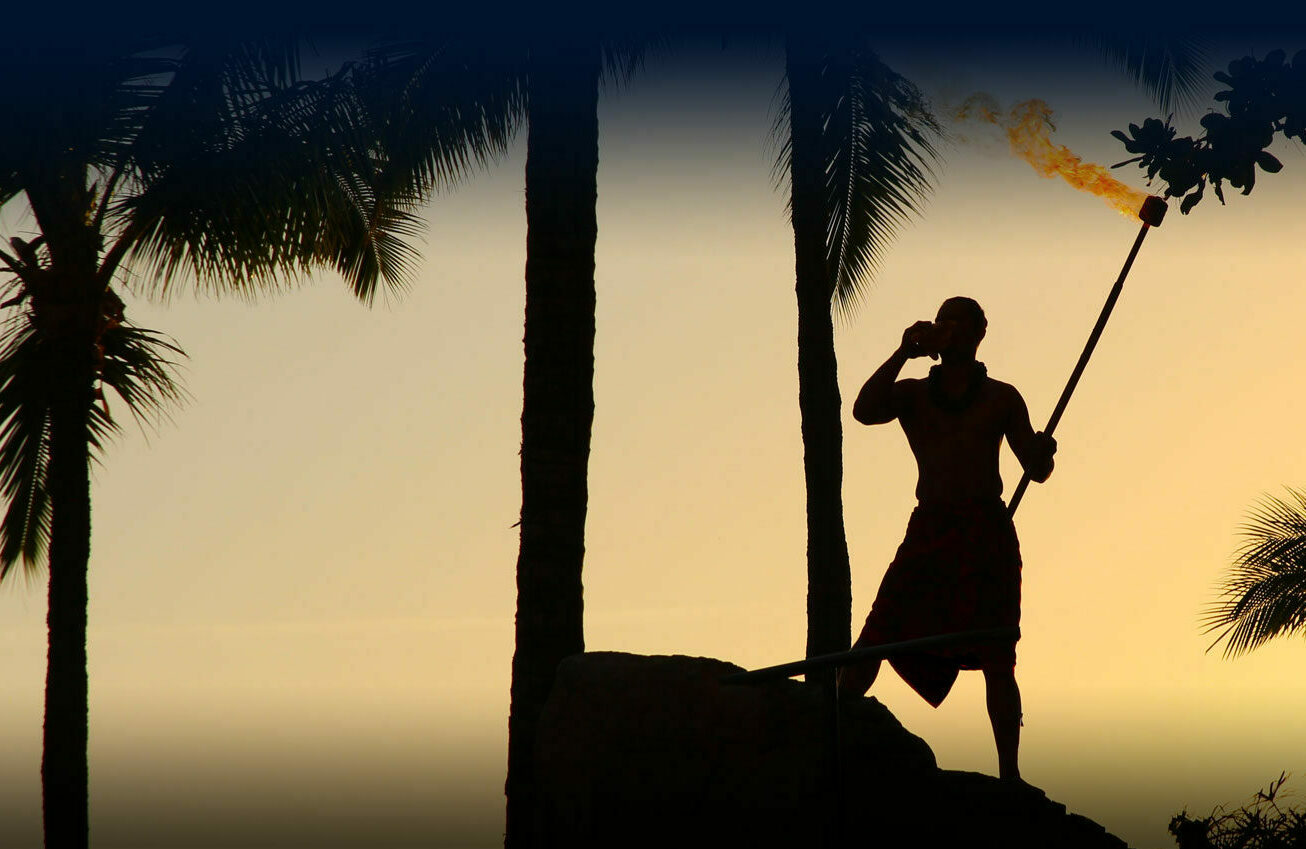 Man stands with a flaming torch in sunset light with palm trees behind him.