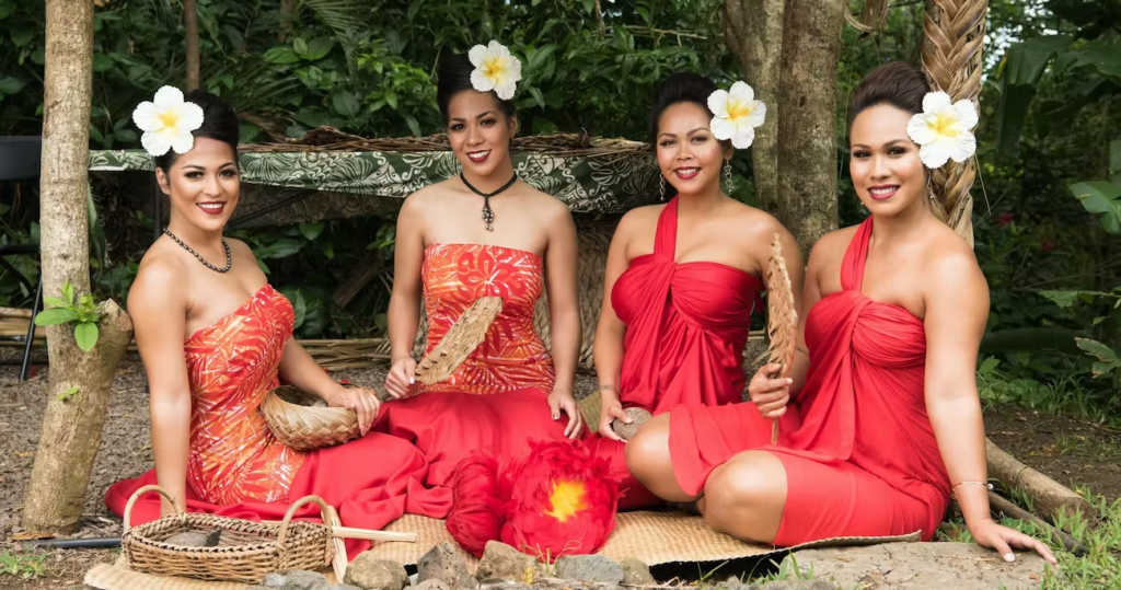 Four hula dancers in red costumes with white flowers in their hair sitting in a row