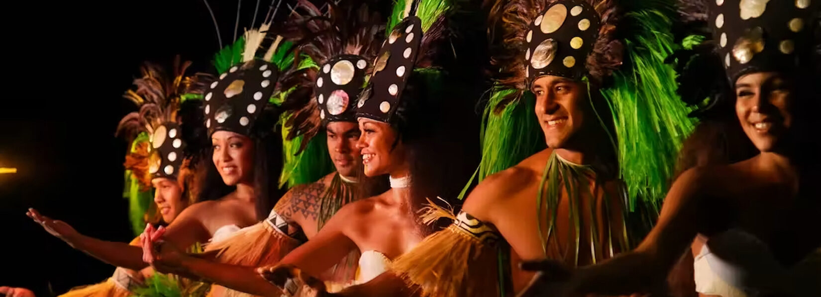 Luau Kalamaku dancers smile on stage in elaborate costumes.