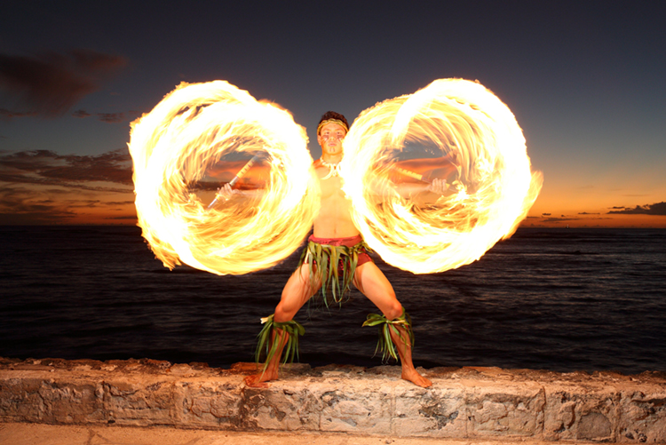 A luau performer spins fire knives in each hand with sunset in the background. 