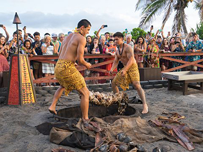 Luau staff remove a whole roasted pig from a traditional cooking pit while guests watch and take photos. 