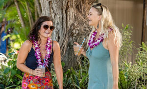 Two ladies wearing flower leis enjoy and drink and a laugh at the luau.