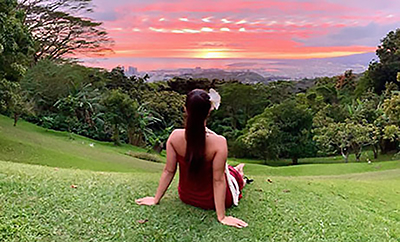 A woman with a flower in her hair watches a colorful sunset over the ocean from a grassy meadow on Oahu. 
