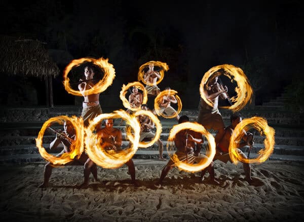 Ten fire knife dancers twirl flaming torches on stage at a luau. 