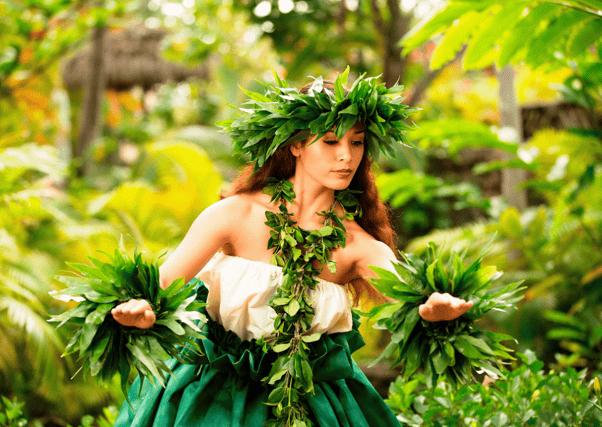 A female hula dancer poses in a green headdress in front of a tropical garden. 