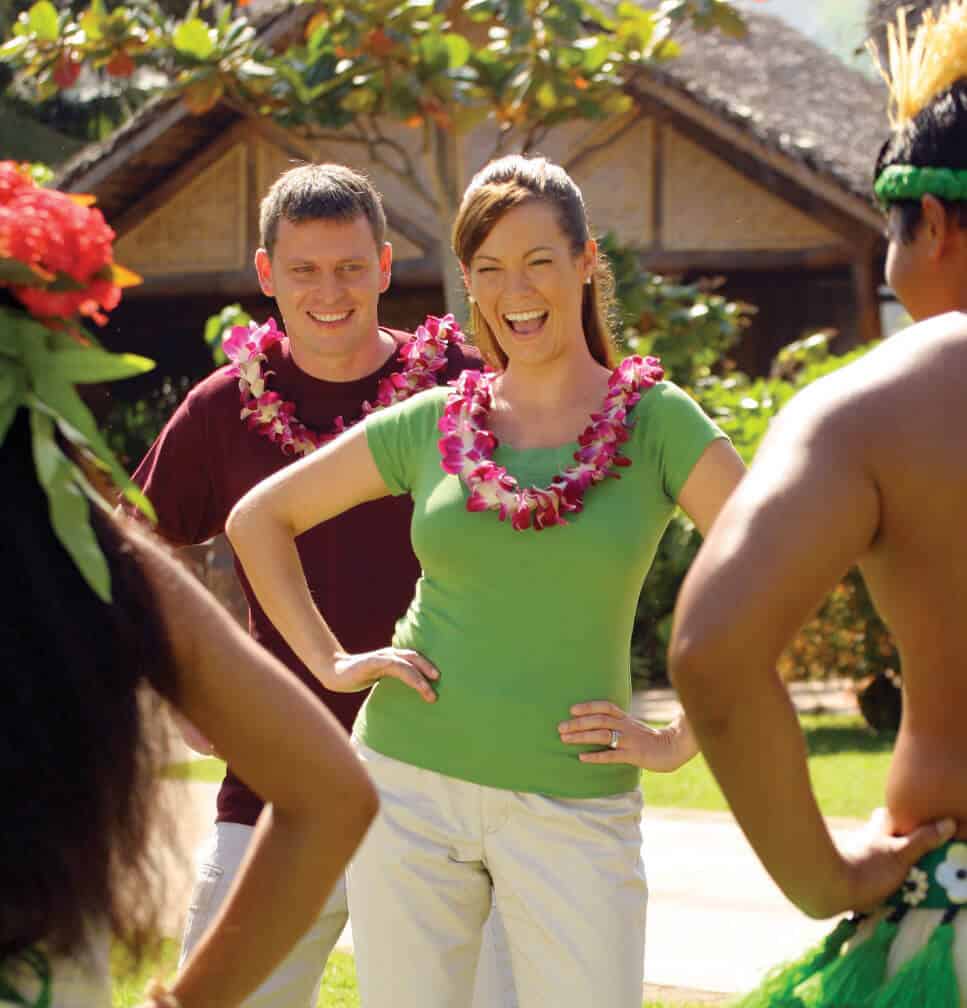 A couple with flower leis learns to hula dance from two luau staff members in Hawaii. 