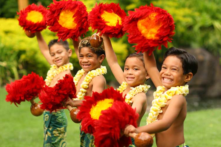 Four children with flower leis and feathered gourds smile for guests arriving at the luau. 