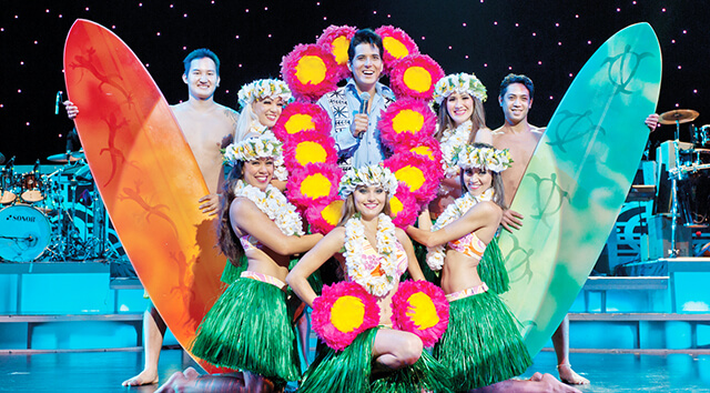 Hula dancers with feathered gourds and surfboards stand near Elvis. 