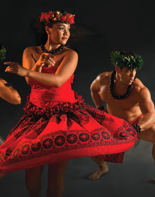 A woman twirls in a red Hawaiian dress with hula dancers in the background.