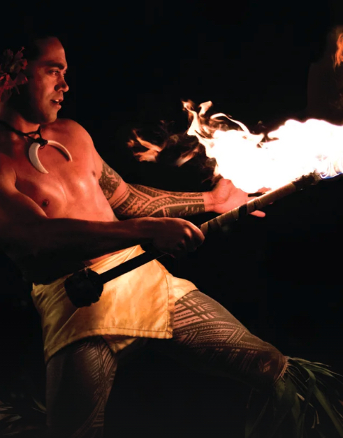 A man with tribal tattoos and boar tusk necklace looks into the flames of a torch. 