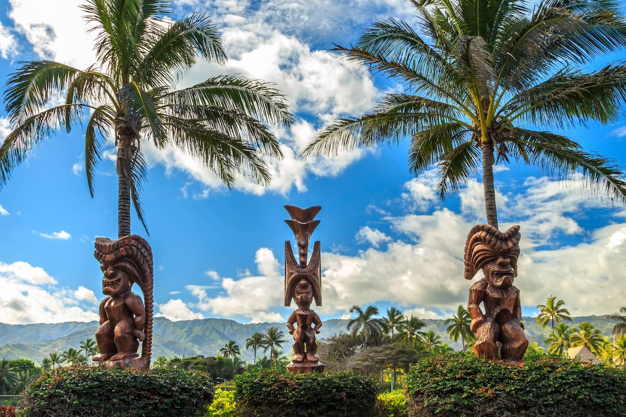 Three Hawaiian tiki statues stand in a tropical garden with mountains in the background. 