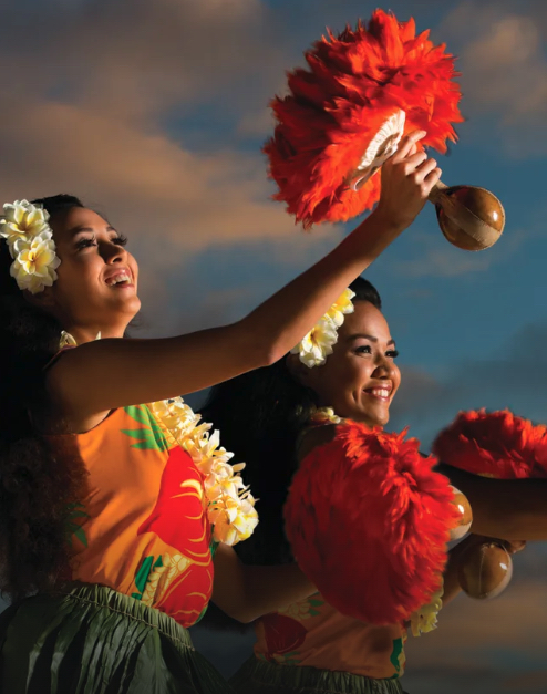 Two ladies in grass skirts and flower leis dance with feathered gourds. 